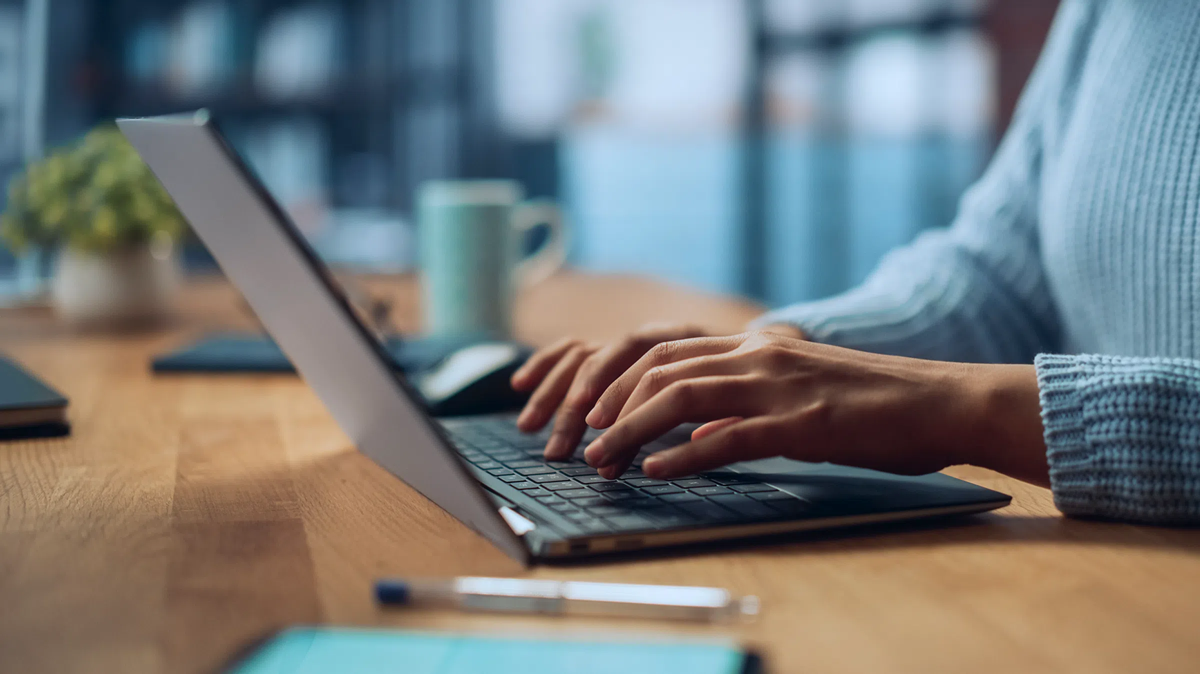 Profissional digitando em laptop em mesa de madeira com café, ambiente corporativo moderno ao fundo desfocado.