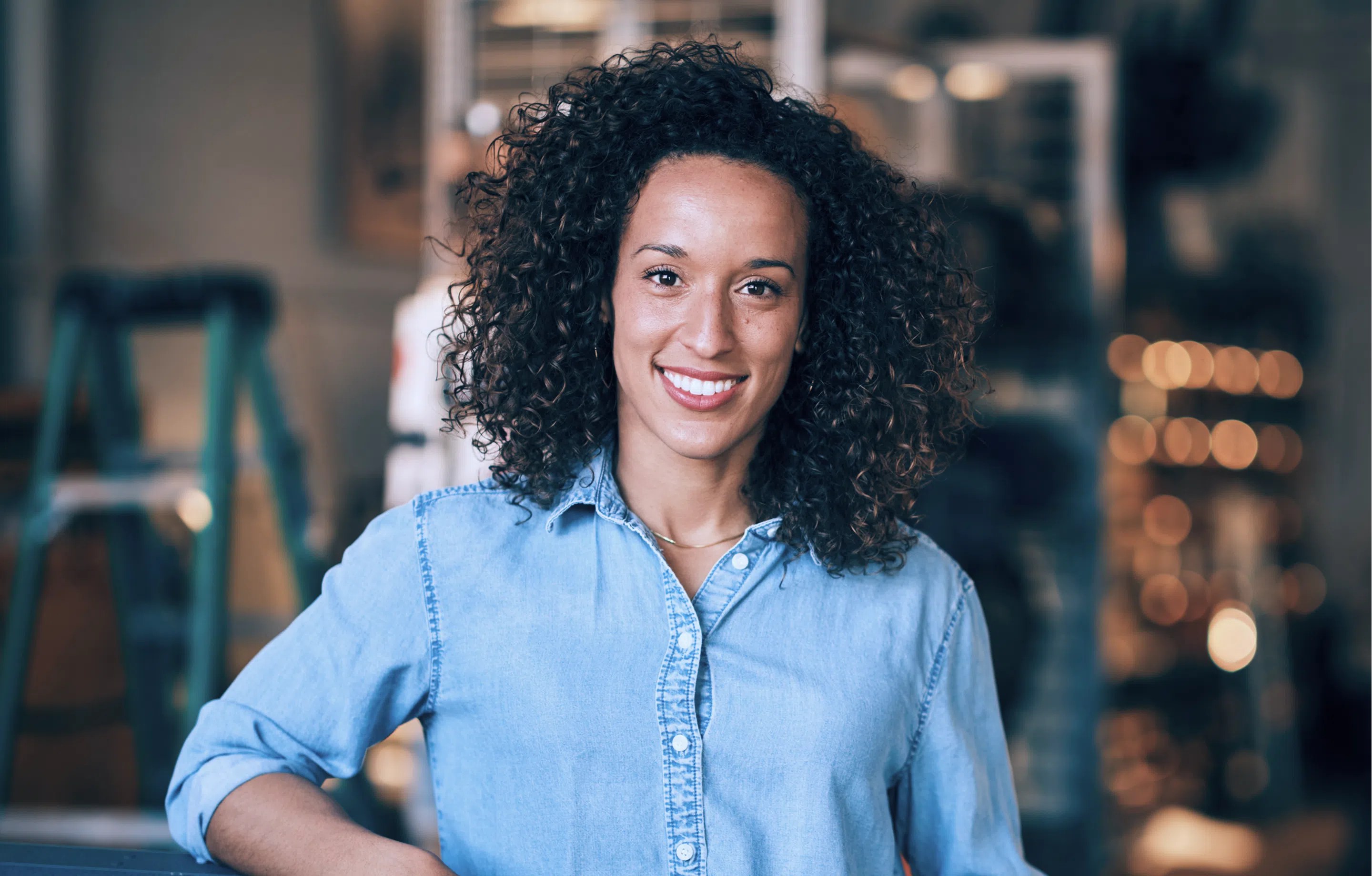 Retrato profissional de mulher sorridente com cabelo cacheado e camisa jeans azul em estúdio com fundo desfocado