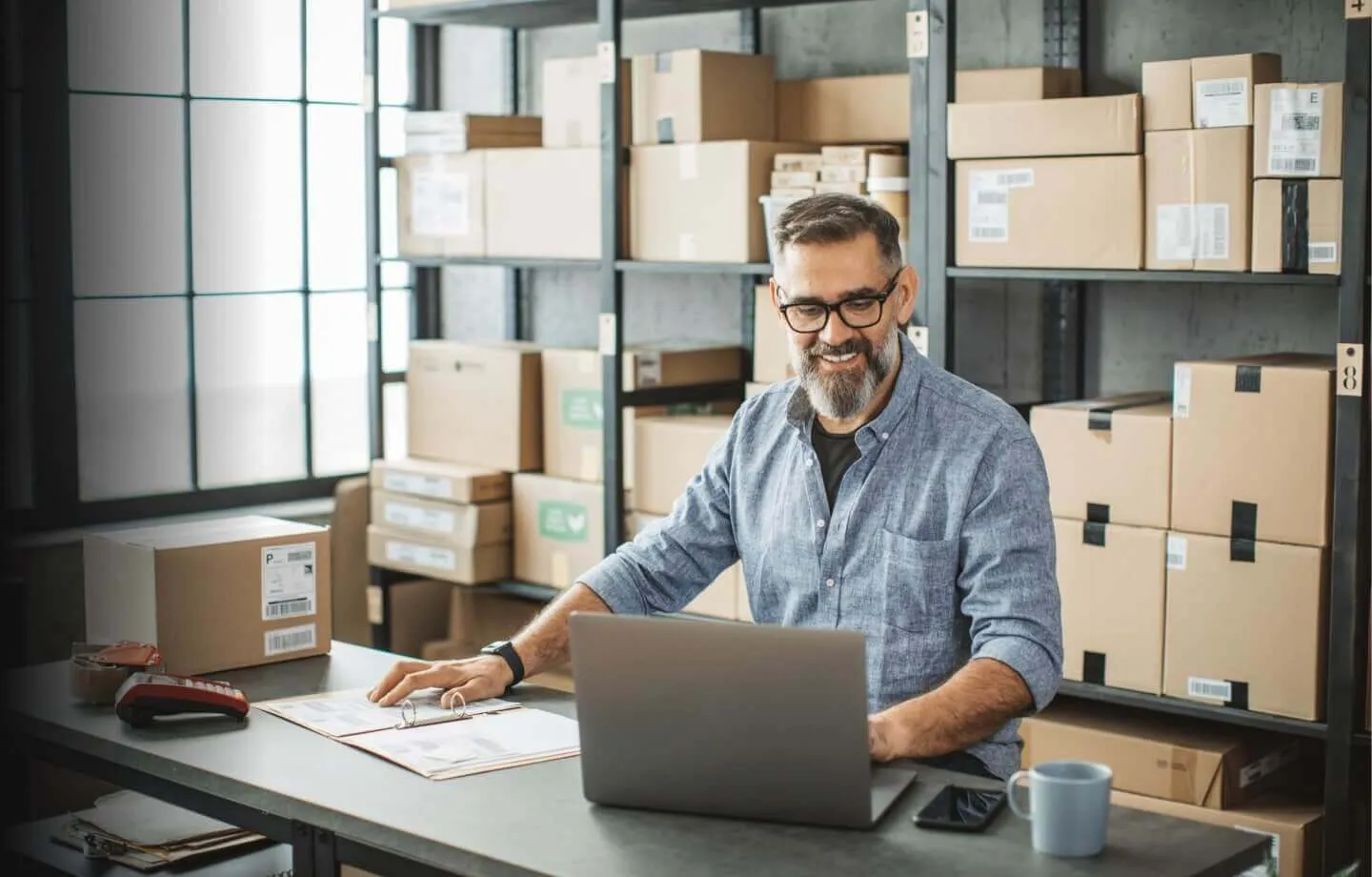 Um homem de negócios, sorrindo, utilizando laptop em ambiente de logística, representando a facilidade e a segurança do desconto de duplicatas do BTG pactual empresas.