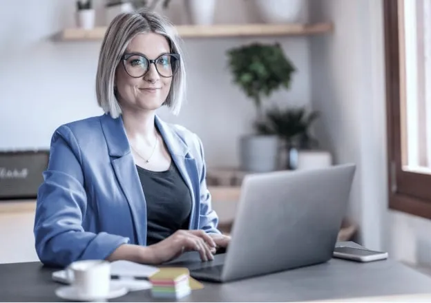 Mulher de óculos e blazer azul trabalhando em um laptop, com plantas e itens de escritório ao fundo.