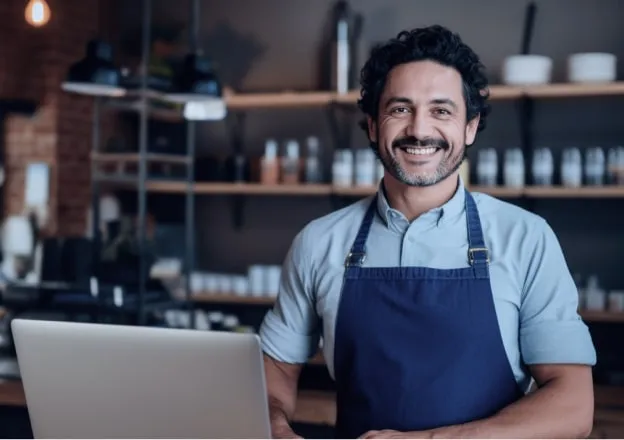 Funcionário com avental, sorrindo e usando um laptop com prateleiras de utensílios ao fundo.