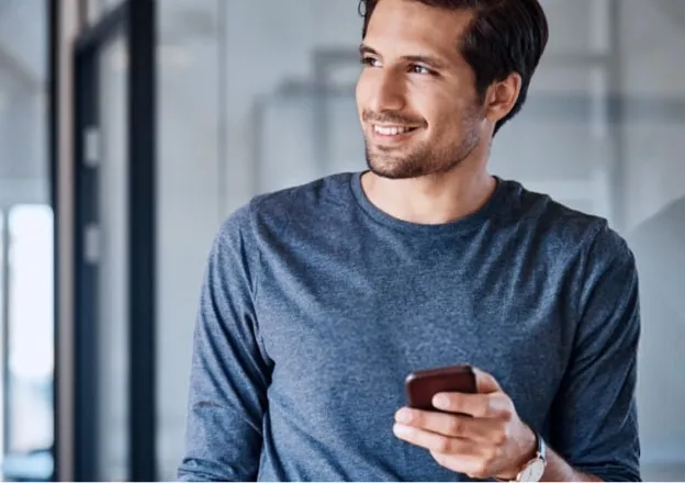 Homem sorridente segurando um smartphone enquanto olha para o lado em um ambiente de escritório.