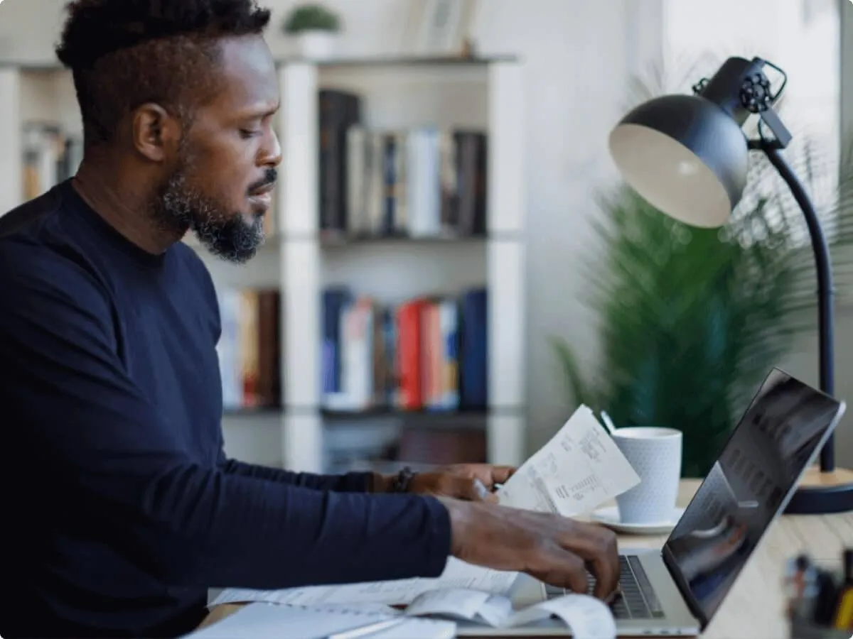 Homem trabalhando em casa, revisando papéis e usando um laptop, com luminária e livros ao fundo.