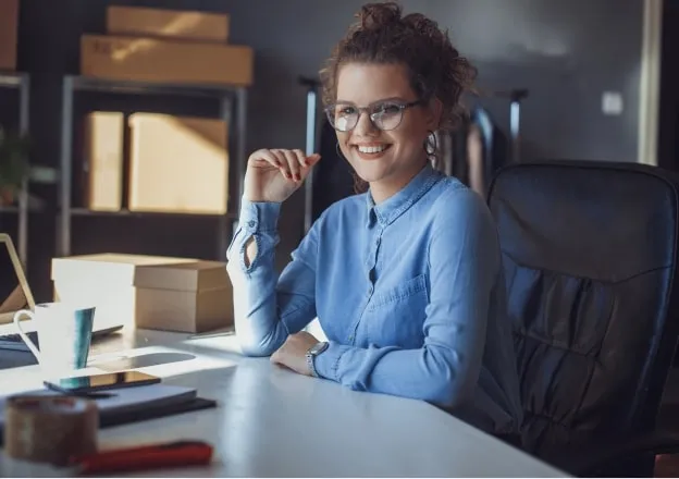 Mulher sorridente usando óculos e camisa azul, sentada à mesa de escritório com caixas ao fundo.