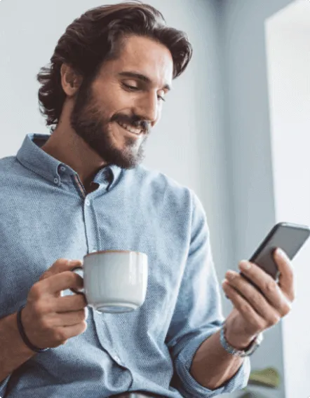 Um homem sorridente, segurando uma xícara de café enquanto utiliza um smartphone, representando a facilidade e conveniência de gerenciar a folha de pagamento empresarial de forma digital com o BTG Pactual Empresas.
