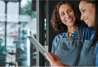 Mulheres em ambiente de trabalho, vestidas com camisas sociais e aventais, sorrindo enquanto organizam o fluxo de caixa da empresa.