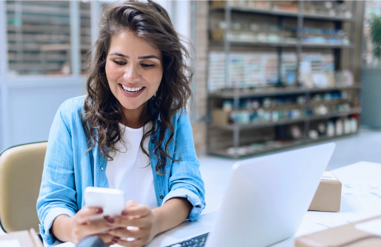 Mulher em um escritório, vestindo camiseta branca e casaco azul, sorrindo enquanto olha para o smartphone.