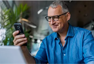 Homem de meia-idade usando camisa azul, sorrindo com confiança para a tela do smartphone exibindo sua conta PJ.