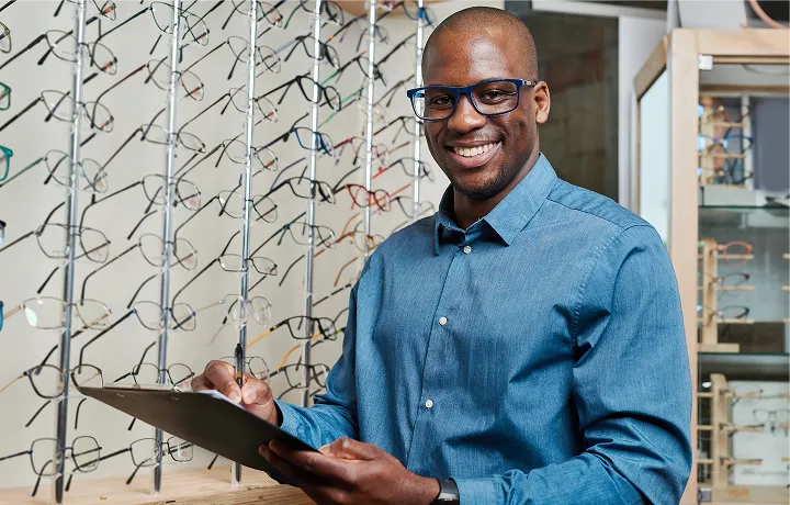 homem negro, sorridente, vestido com uma camisa azul. Seu fundo tem um mostruário com lentes de óculos.