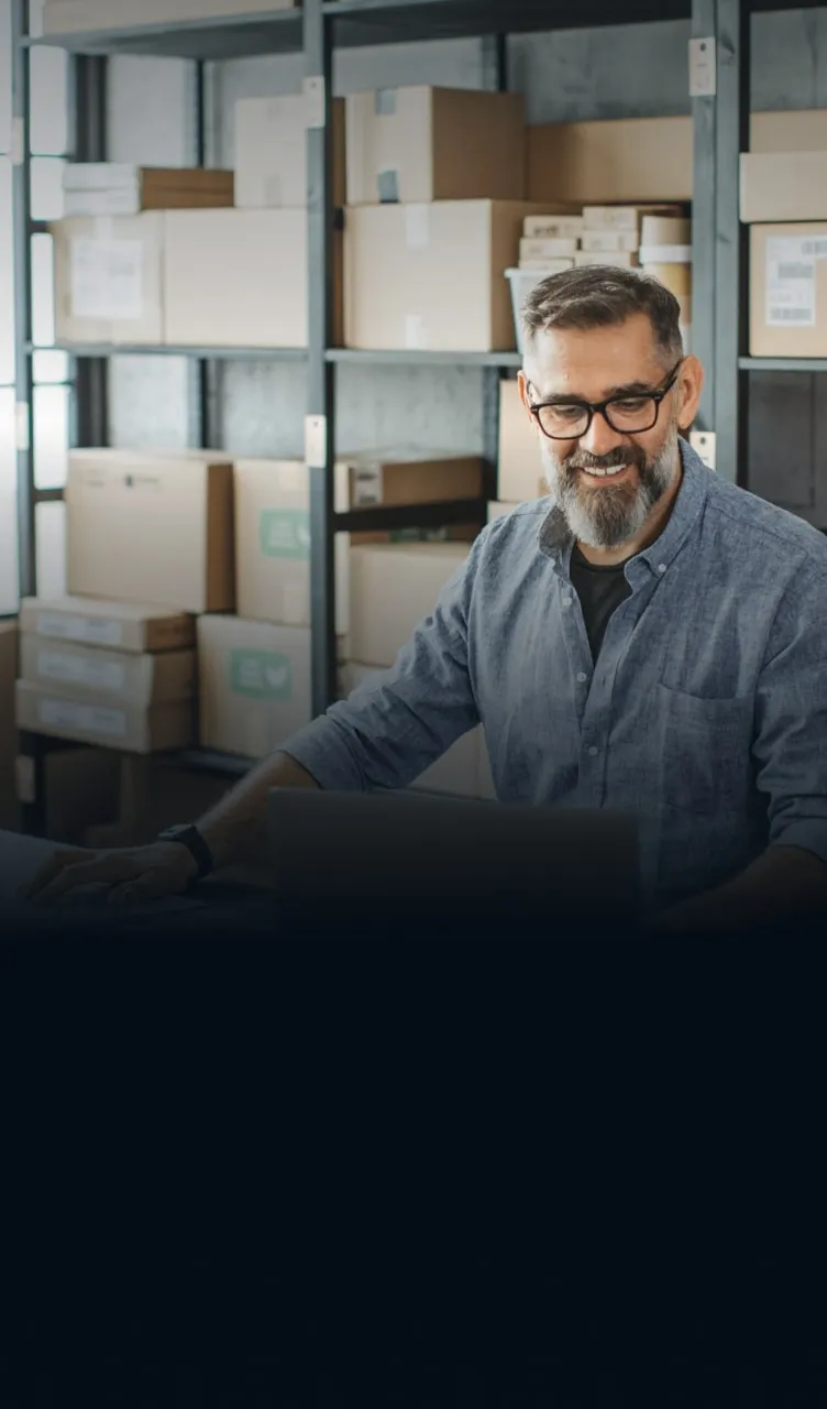 Um homem de negócios, sorrindo, utilizando laptop em ambiente de logística, representando a facilidade e a segurança do desconto de duplicatas do BTG pactual empresas.