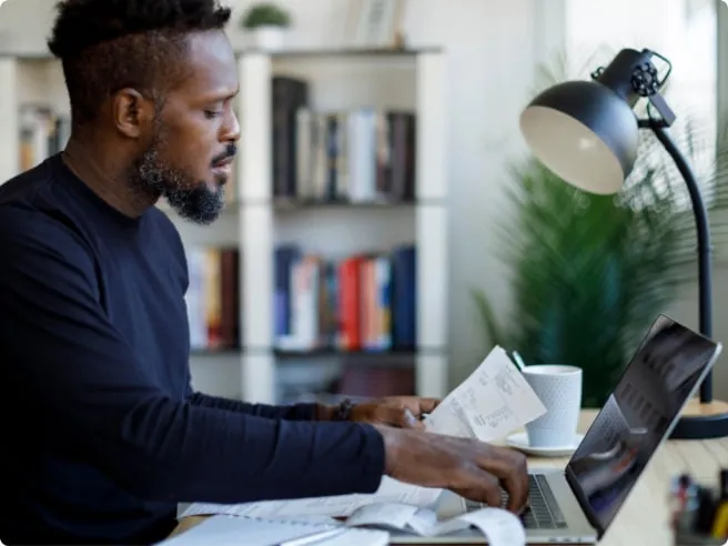 Homem trabalhando em casa, revisando papéis e usando um laptop, com luminária e livros ao fundo.