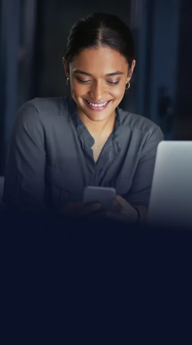 Empresária sorridente utilizando seu smartphone enquanto trabalha no laptop, representando a praticidade e a segurança do cheque especial empresarial BTG Pactual