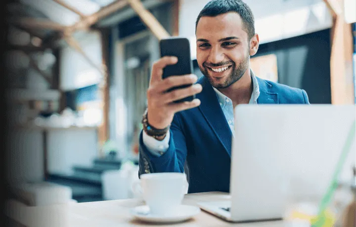 Homem sorridente utilizando um smartphone enquanto trabalha em um laptop, destacando a praticidade e modernidade na gestão de folha de pagamento empresarial oferecida pelo BTG Pactual Empresas.