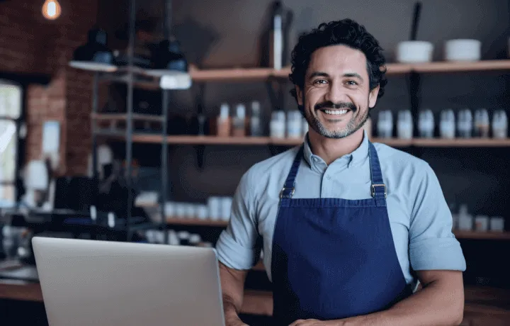 Homem usando avental azul e sorrindo em frente a um Notebook por poder antecipar os recebíveis da empresa