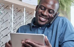 homem sorrindo, falando ao celular e olhando para um tablet.